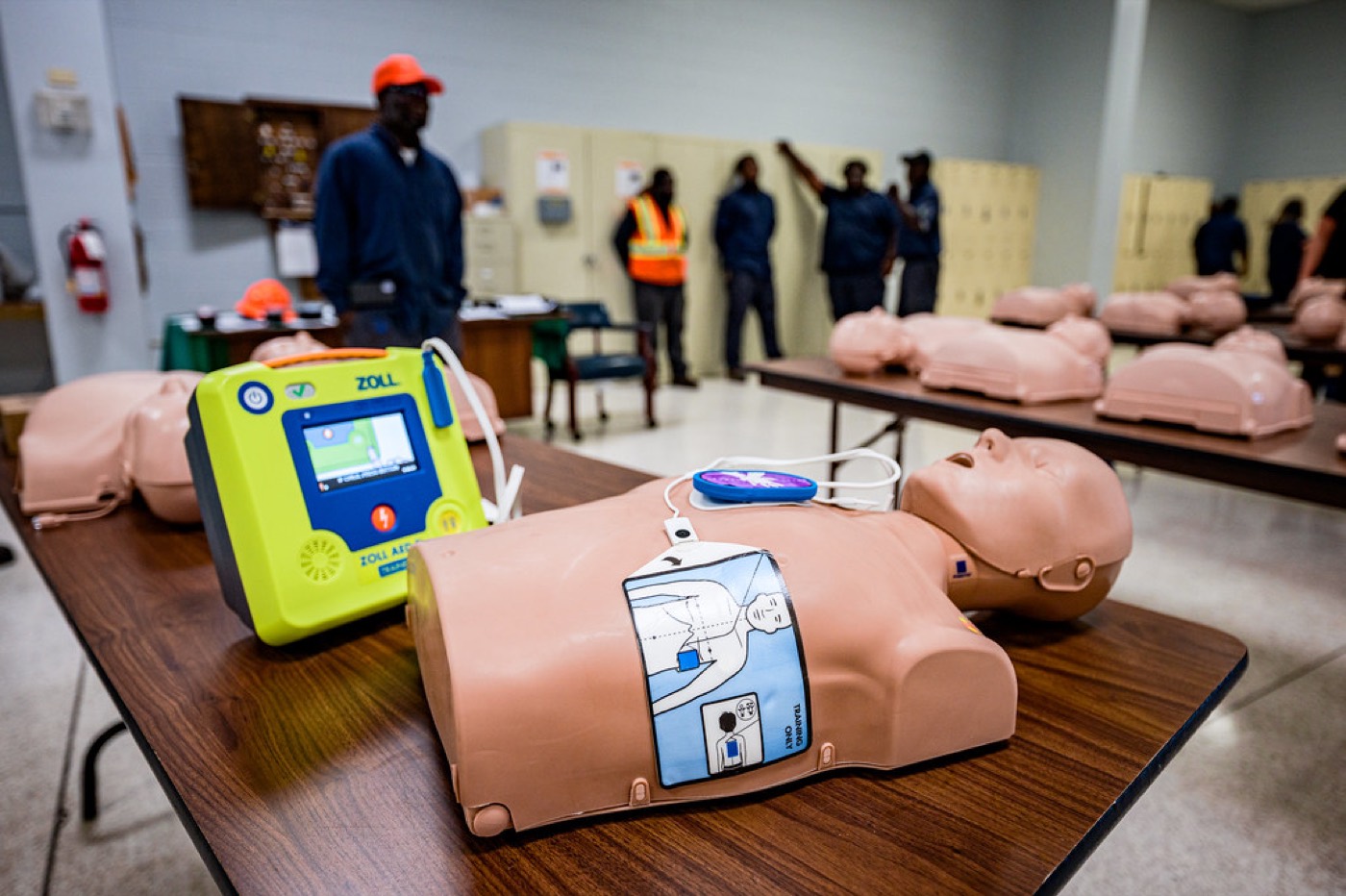 An AED trainer and CPR manikin set up on a table in a workplace, with staff training in the background.