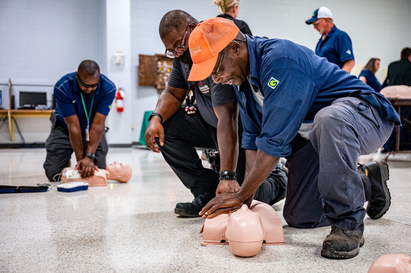 Students kneeling on the floor performing chest compressions on manikins while a trainer coaches them.